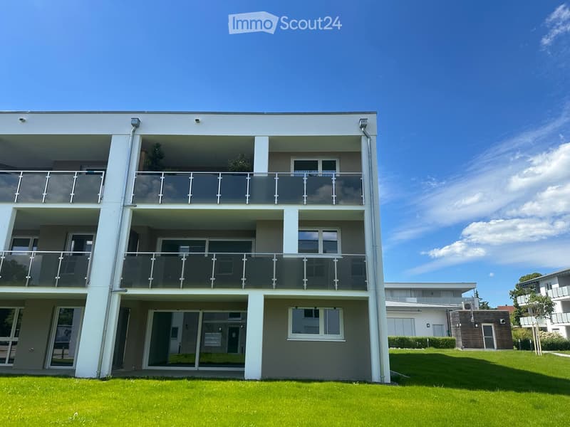 Modern two-story building with large glass balconies and white exterior walls, surrounded by green lawn.