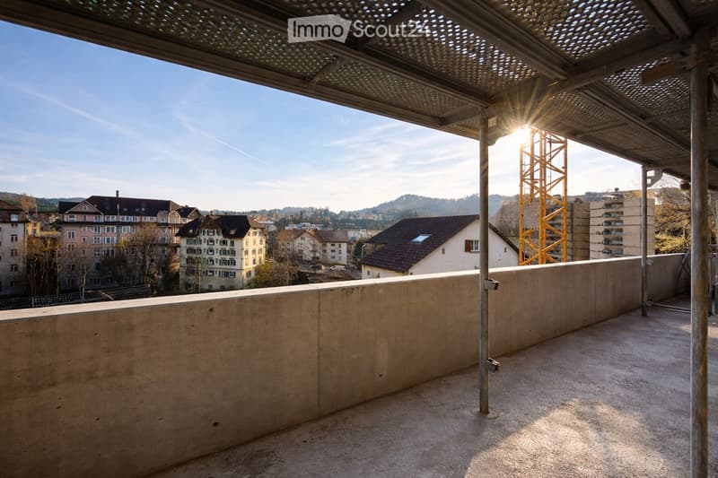 Empty balcony with concrete floor and low wall, metal railing, and a perforated metal roof providing partial shade. The view shows a cityscape with residential buildings and a construction crane in the distance.