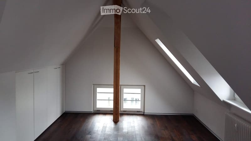 Unfurnished Room. An empty attic room with wooden flooring, white walls, and large windows allowing natural light to enter. The room features a slanted ceiling with skylights and a central wooden beam.