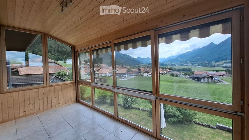 Bright, wooden-framed room with large windows offering panoramic views of a village and mountains. The room features a tiled floor and exposed wooden beams on the ceiling.