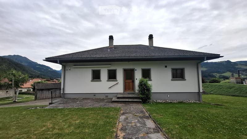 Single-story house with white exterior, wooden front door, and two chimneys. Surrounded by a grassy lawn and pathway leading to the entrance, with mountains in the background.