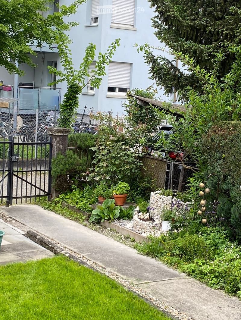 Garden area with a concrete pathway, potted plants, shrubs, and a small stone fountain. A black metal gate leads to a fenced area with bicycles and a light blue building in the background.