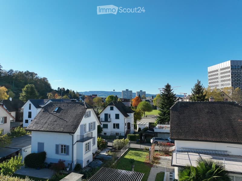 Aerial view of a residential neighborhood with multiple houses, gardens, and a clear blue sky. The houses have pitched roofs, balconies, and are surrounded by greenery and trees.