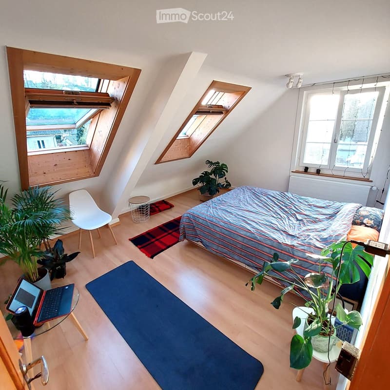 Cozy attic bedroom with wooden floors, large windows, skylights, bed with striped bedding, laptop on a small table, potted plants, and a blue yoga mat.