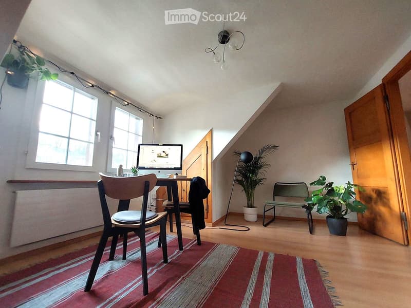 Cozy attic room with wooden floors, large windows, a desk with a computer, a chair, a bench, and potted plants.