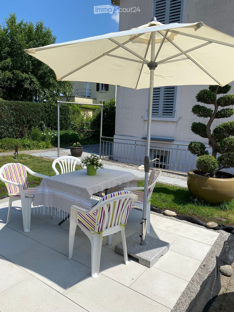 Outdoor patio area with a table, chairs, umbrella, and potted plants, surrounded by a well-maintained garden and a building in the background.