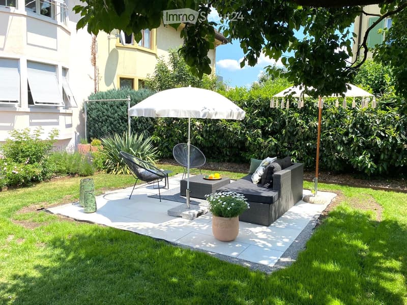 Well-maintained garden with a patio area featuring a sofa, chairs, a table, and an umbrella, surrounded by greenery and a potted plant.