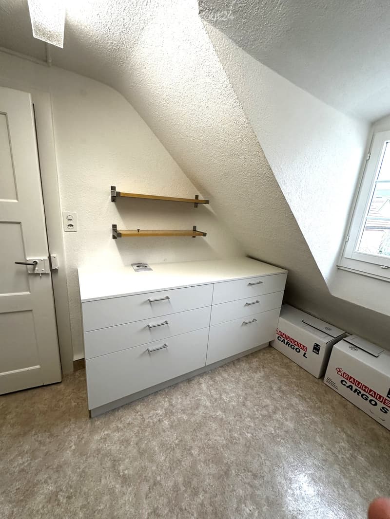 An attic room with white walls, a slanted ceiling, and a carpeted floor. Features a white cabinet with multiple drawers and two wooden shelves mounted on the wall. There are two cardboard boxes on the floor near the window.
