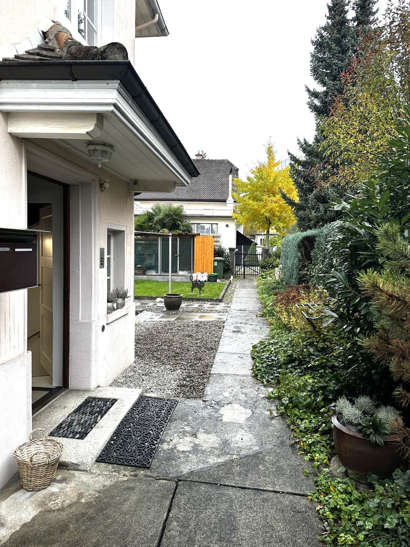 A narrow garden pathway lined with potted plants and shrubs, leading to a house entrance. The pathway is paved with concrete slabs and features decorative metal grates near the entrance. The house has a white facade with a small porch area and a basket pl