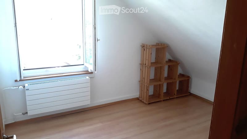 Empty room with wooden flooring, white walls, a window with a radiator below, and a wooden shelf unit in the corner.