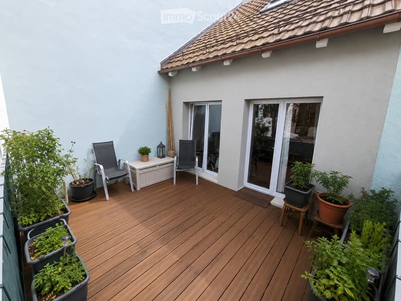 Wooden deck terrace with two chairs, a small table, potted plants, and a lantern. The terrace has a slanted roof and overlooks a building entrance.