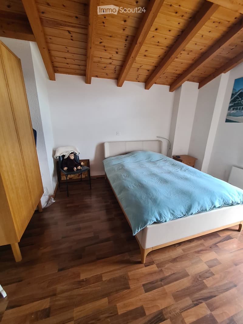 Cozy bedroom with wooden floors, slanted wooden ceiling, white walls, a bed with light blue bedding, a small nightstand, and a chair with a stuffed animal on it.