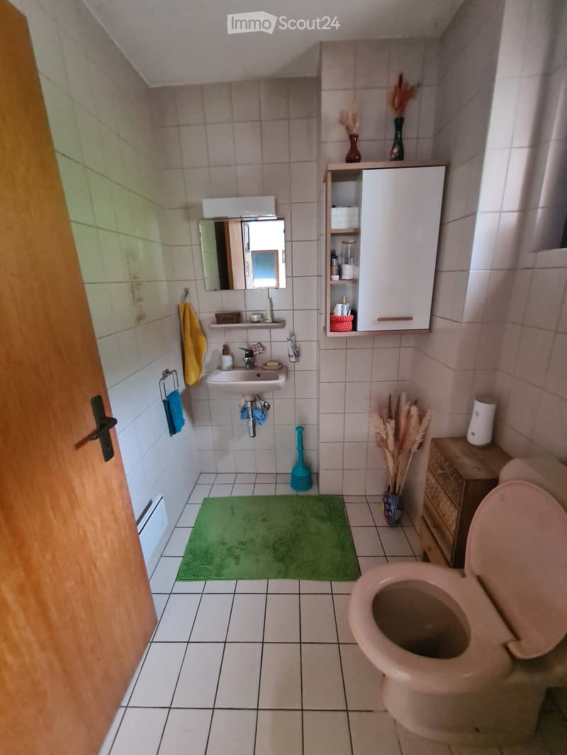 Small bathroom with white tiled walls and floor, featuring a toilet, sink with mirror, cabinet, towel, vase with dried plants, and a green rug.