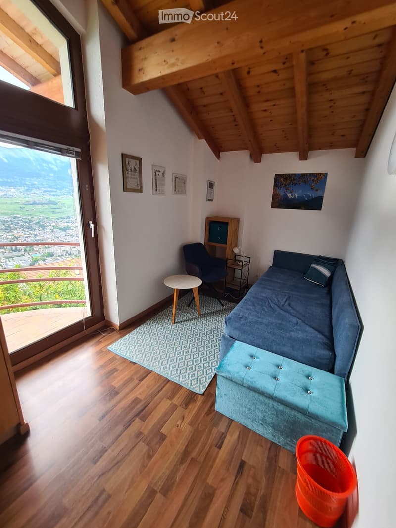 Cozy attic bedroom with wooden floors, a blue sofa, a small round table, a chair, a lamp, and a view of the city from a large window.