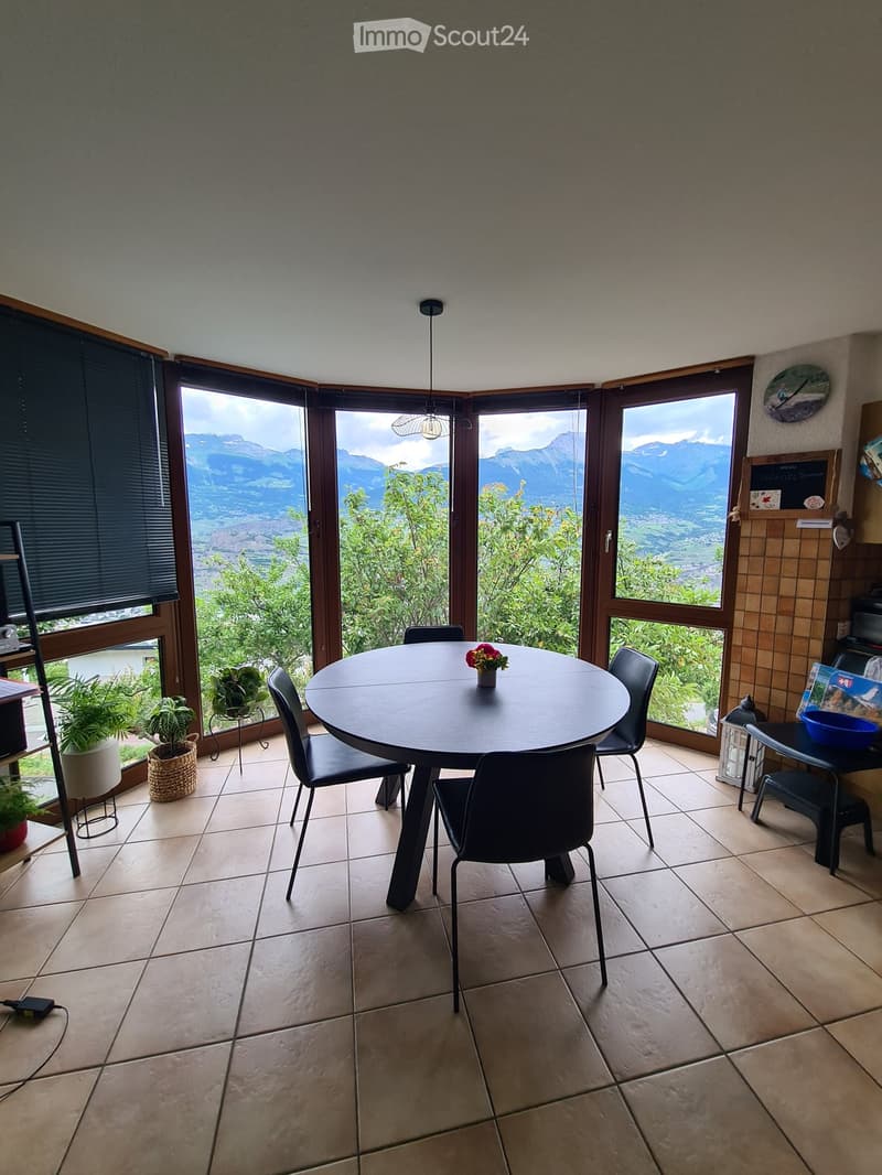 Bright dining area with large windows offering a scenic mountain view, round table with chairs, potted plants, and decorative items.