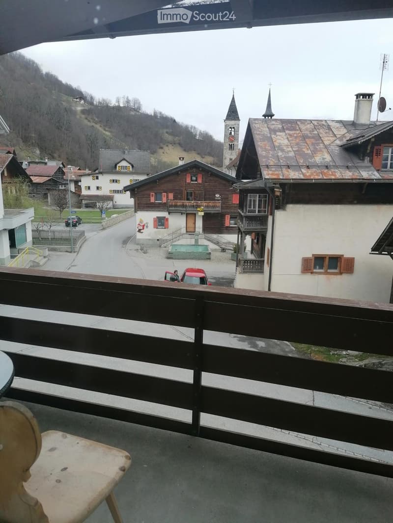 Balcony overlooking a village with traditional wooden houses, a church tower, and a small fountain in the foreground.