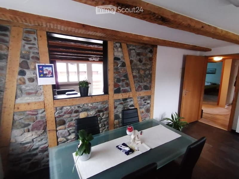 Dining area with a glass table, chairs, and a small plant, adjacent to a stone-walled room with a bed and window.