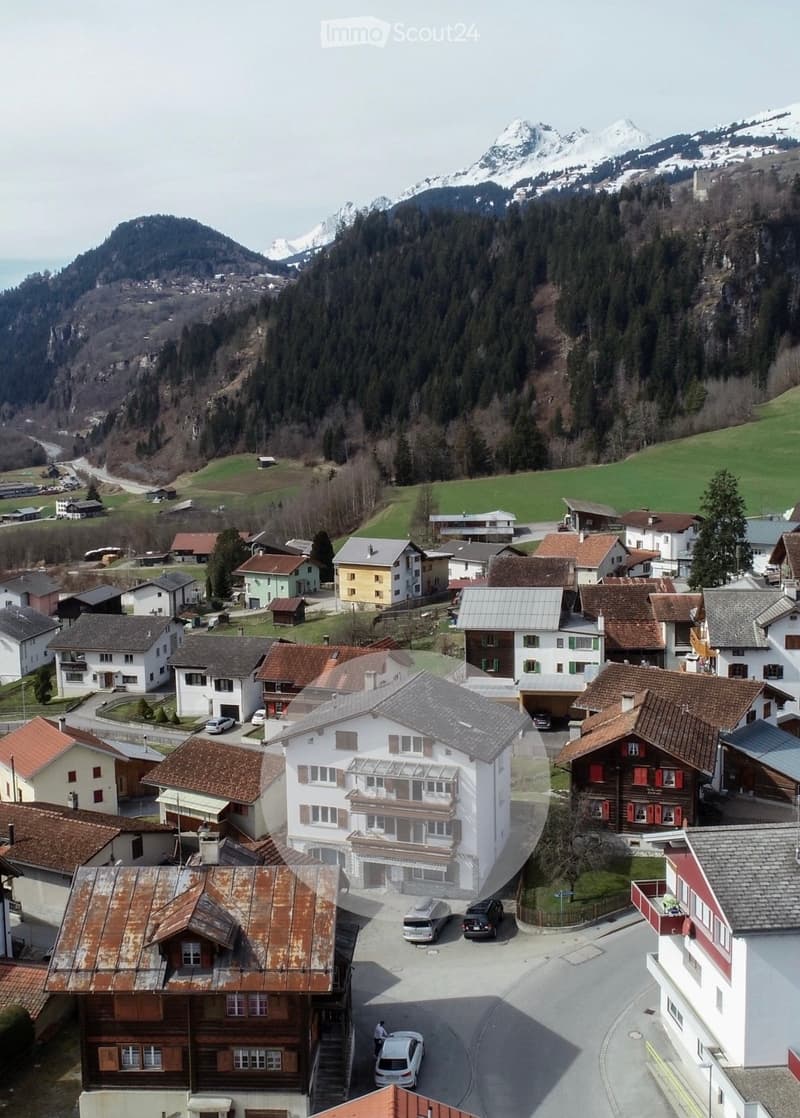 Aerial view of a small village nestled in a valley with surrounding mountains. The village features a mix of residential houses with varied roof styles, including red-tiled and grey roofs, and some buildings have visible balconies. The area includes roads