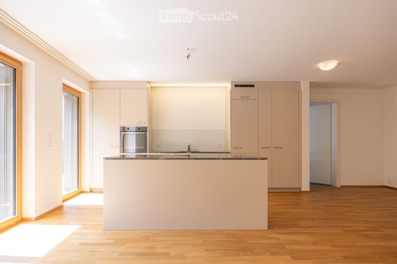 Modern kitchen with light-colored cabinets, a central island, and a built-in oven. The space features wooden flooring and large windows allowing natural light to fill the room.