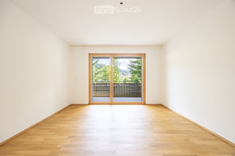 Empty room with light wooden flooring and white walls, featuring a large sliding glass door leading to a balcony with a view of greenery and mountains.