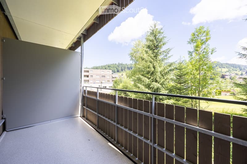 Balcony with wooden and metal railing, overlooking a green landscape with trees and distant buildings.