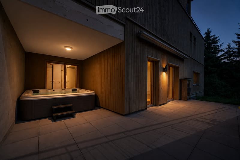 Outdoor patio area with a hot tub, wooden decking, and steps leading to the tub, illuminated by warm lighting.