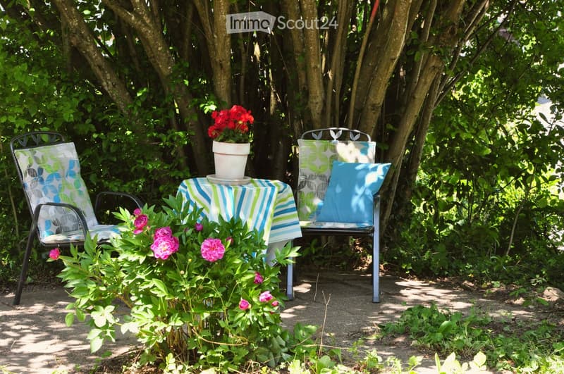 A serene garden setting with a small table covered in a blue and white striped cloth, surrounded by lush greenery and blooming flowers, including pink roses and red geraniums. Two metal chairs with colorful cushions are placed around the table, creating a