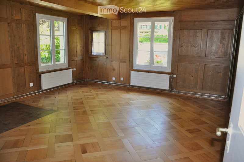 Empty room with wooden panel walls, herringbone wooden flooring, and large windows with radiators beneath them.