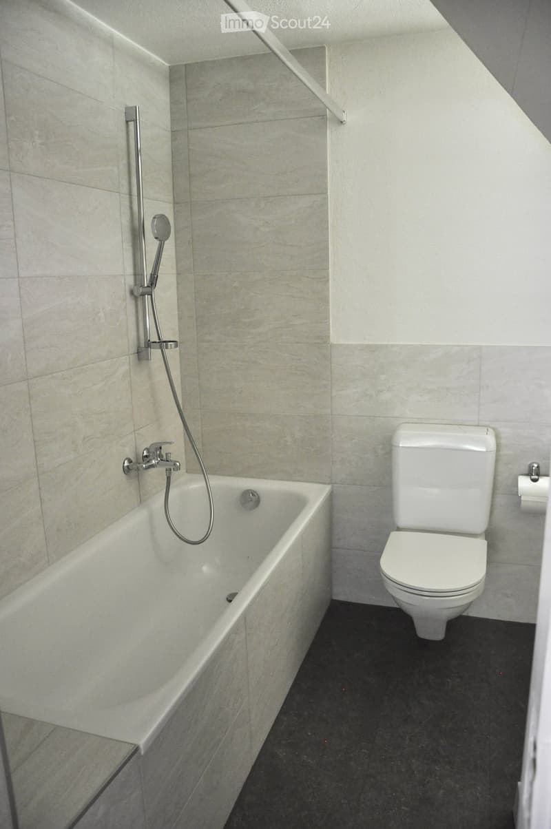 Modern bathroom with a white bathtub, toilet, and showerhead, featuring beige tiled walls and a dark floor.