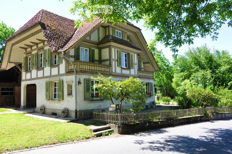 Two-story house with a tiled roof, green shutters, and a balcony on the upper floor. The house features a wooden fence, a small garden, and a driveway.