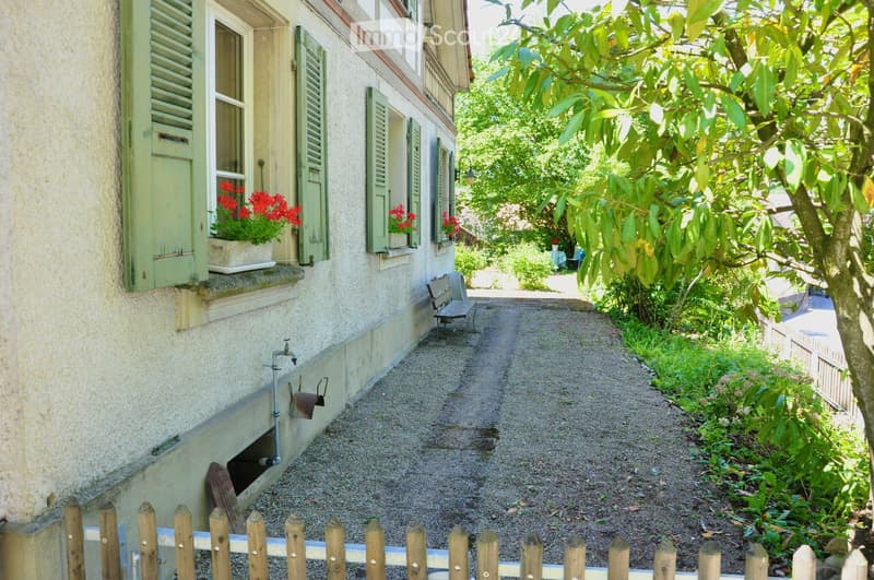 A narrow gravel pathway leads to a house with light green shutters and flower boxes. The pathway is bordered by a wooden fence and lush greenery.