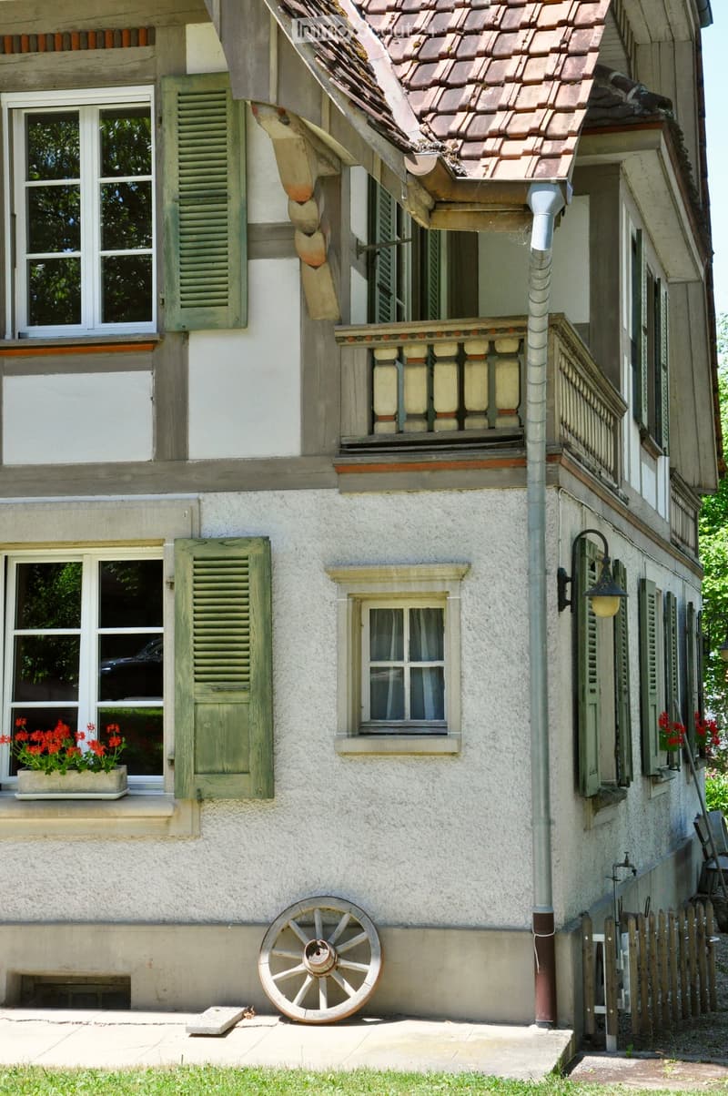 Two-story house with white plaster walls, green shutters, and a tiled roof. Features include a balcony with ornate railings, a decorative wooden wheel on the ground, and flower boxes with red flowers on the windowsills.