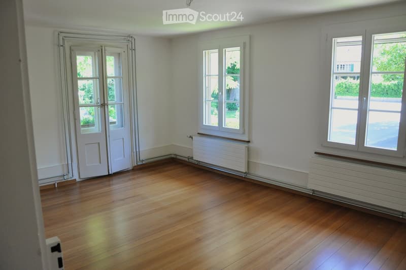 Empty room with wooden flooring, white walls, and large windows. Radiators are visible along the walls, and a glass door leads to an outdoor area.