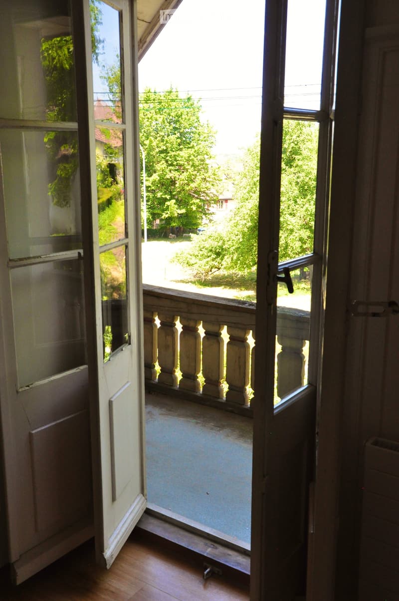 Open balcony with wooden balustrade, overlooking a green, tree-filled area with a road and houses in the background.