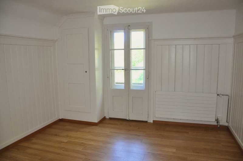 Empty room with wooden flooring, white walls, a radiator, and a large window with double doors.