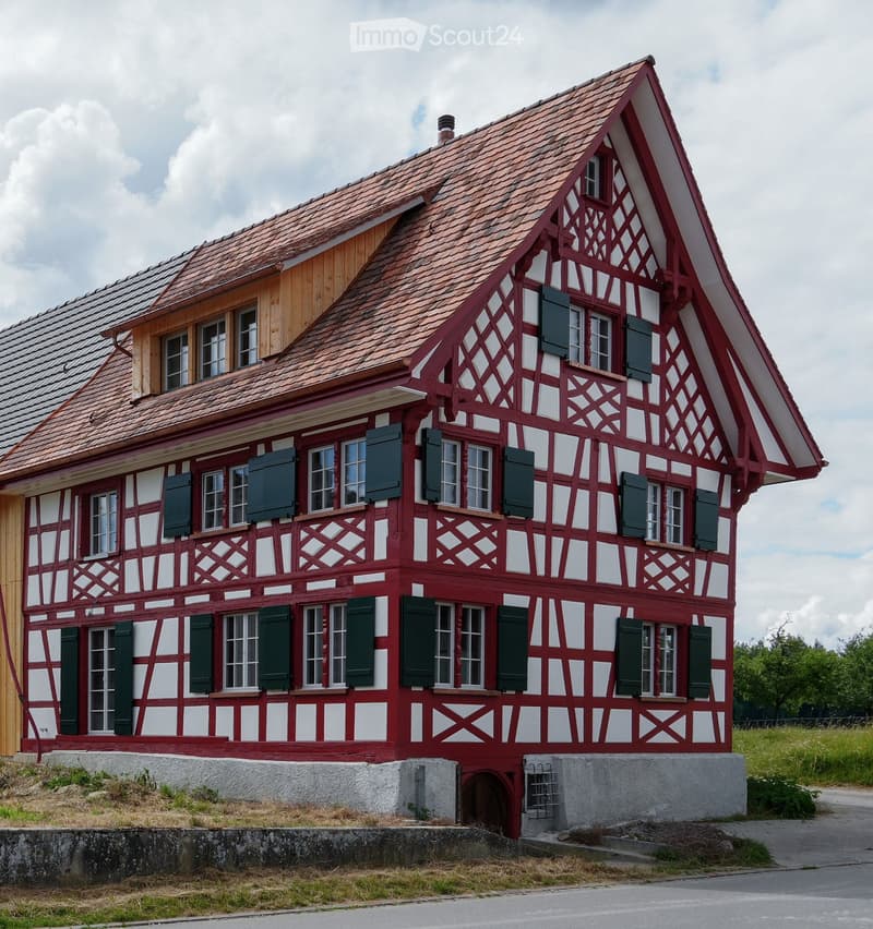 Traditional red and white half-timbered house with multiple windows and green shutters, situated on a raised ground floor with a stone base.
