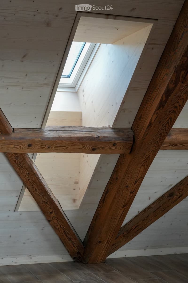 An empty room with exposed wooden beams and a skylight window, featuring a wooden floor and white paneled walls.