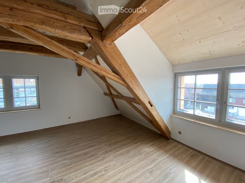 Empty attic space with wooden beams, wooden flooring, and large windows offering a view of the neighborhood.