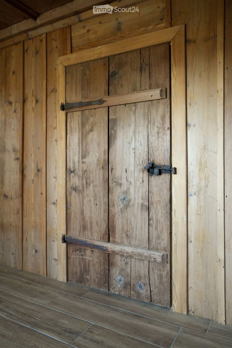 A wooden hallway with a large, rustic wooden door featuring metal hinges and a latch, set in wooden paneled walls and a wooden floor.