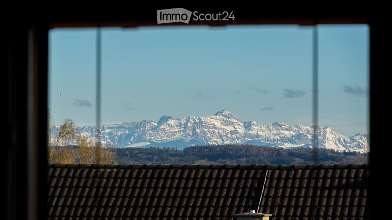 View from a balcony or terrace showing a clear blue sky and snow-capped mountain range in the distance, with a tiled roof visible in the foreground.