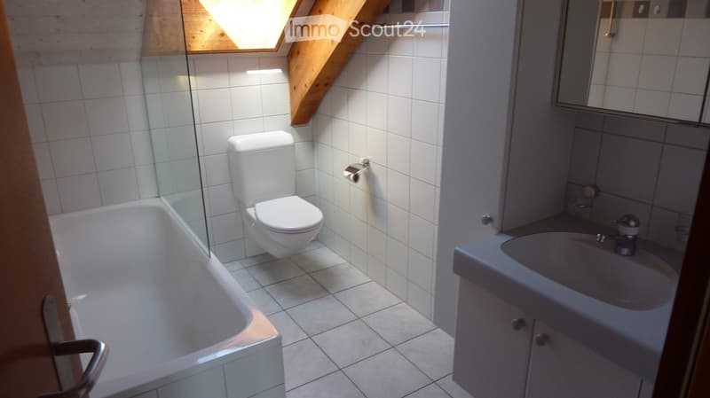 Modern bathroom with white tiles, a bathtub, toilet, sink, and a mirror above the sink. The room features a skylight window and wooden beams on the ceiling.