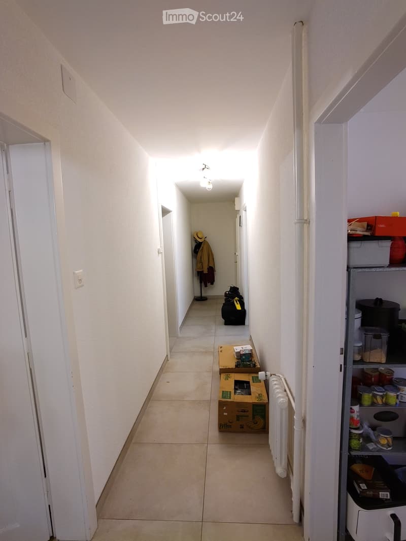 A narrow hallway with tiled flooring, white walls, and several doors. Visible items include a coat rack with jackets, a suitcase, and some boxes on the floor.