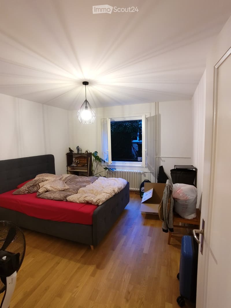A cozy bedroom with a wooden floor, a large bed with a red cover, a nightstand, a fan, a chair, and a radiator near the window.