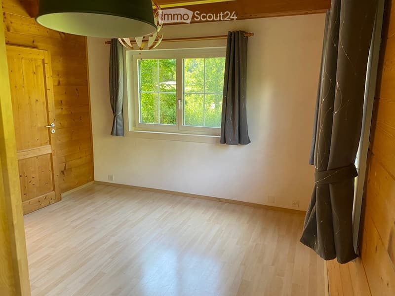 Empty room with wooden flooring, white walls, large window with dark curtains, and a wooden door.