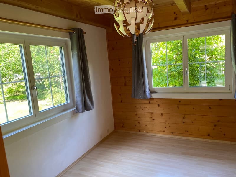 Empty room with wooden walls and flooring, large windows with grey curtains, and a modern chandelier.