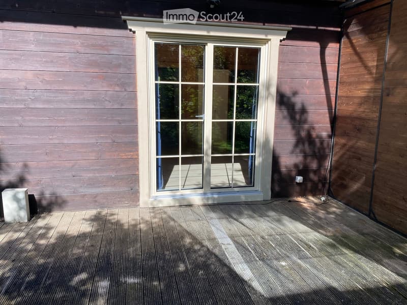 Wooden deck with a large glass door leading to the interior, surrounded by wooden walls and shadows from nearby trees.