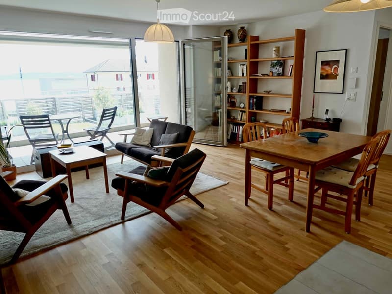 Bright living room with wooden flooring, large glass sliding doors leading to a balcony, a sofa, coffee table, dining table with chairs, bookshelf, and decorative items.