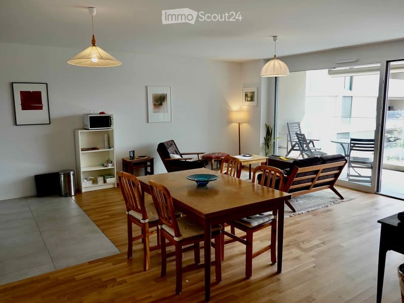 Bright, open living and dining area with wooden flooring, large windows, and a balcony. Includes a dining table with chairs, a sofa, a small table, a shelf with a microwave, and decorative items.