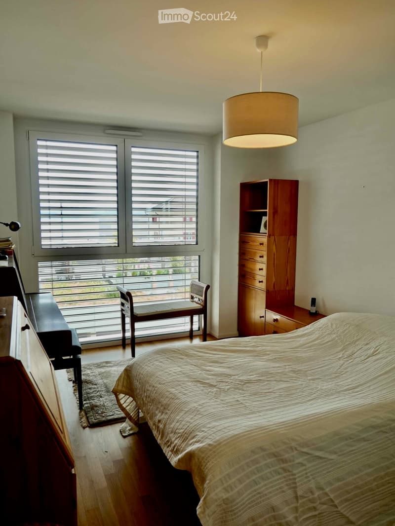 Bright bedroom with wooden floor, large window with blinds, bed with beige bedding, wooden wardrobe, chair, and a small table near the window.