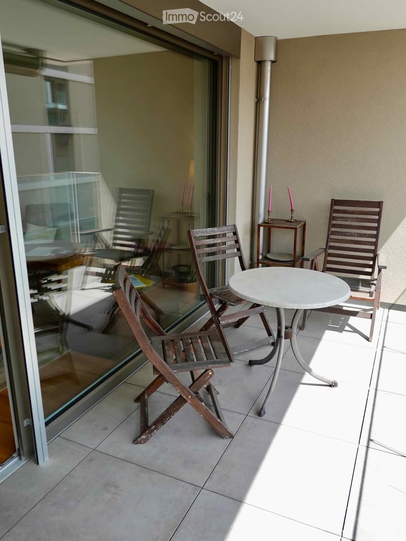 Balcony with tiled floor, white round table, four wooden chairs, small wooden stool with candles, and a view of the interior room through large glass doors.